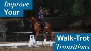 A person riding a horse in an outdoor arena demonstrates a smooth walk-trot transition. Text on the image reads Improve Your Walk-Trot Transitions. Trees, a fence, and a dressage letter S are visible in the background.