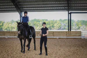 A rider demonstrating classical dressage alignment is mounted on a dark bay horse inside a covered arena. An instructor stands at the quarter line, offering guidance and encouragement with a smile. Lush trees are visible beyond the open sides of the arena, providing natural light and scenery.