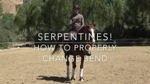 A person wearing a helmet sits on a brown horse in a sandy outdoor arena with trees in the background. Text on the image reads: Serpentines! How to properly use bending aids in horse riding to change bend.
