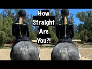 Two people in black riding outfits and helmets sit on gray horses, viewed from behind, with the text How Straight Are You?! in the center—highlighting rider position and symmetry against a backdrop of trees and a riding arena.