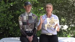 Two women sit outdoors on a bench. One gestures while holding a human pelvic bone model, explaining the muscles you use when you ride; the other listens with her hands folded in her lap. Trees are in the background.