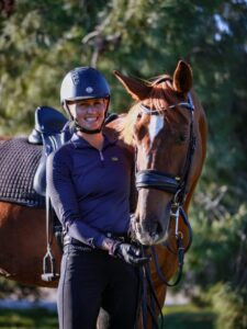 A smiling person in a riding helmet and gear stands outdoors, holding the reins of a brown horse with a black saddle, ready for Dressage Training against a backdrop of green trees.
