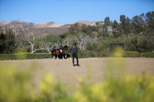 A person on horseback is being observed by another during dressage training in an outdoor riding arena, surrounded by greenery and mountains under a clear blue sky.