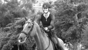 A person wearing a helmet and riding gear smiles while practicing dressage training on a horse outdoors, surrounded by trees. The image is in black and white.