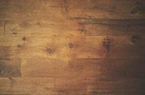 Close-up of a wooden surface with a warm, brown tone. The wood grain and texture are visible, with some natural knots and color variations throughout the planks.