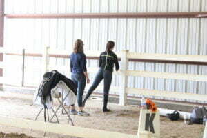 Two women stand by a fenced riding arena, one stretching her leg on the fence to work on rider fitness. A saddle and riding gear sit on a nearby rack, and a dressage marker with flowers is in the foreground. Sign up for our FREE Mini Course!.