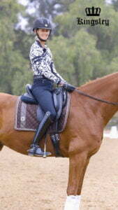 A smiling equestrian wearing a helmet, patterned long-sleeve shirt, riding pants, and Kingsley Boots sits on a chestnut horse with a brown saddle pad. The word Kingsley with a crown logo appears in the upper right corner.
