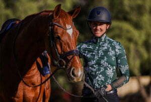 A smiling person in a green floral shirt and black helmet stands beside a brown horse, gently holding its bridle. They are outdoors amidst greenery, ready for groundwork training in a serene setting.