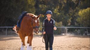 A smiling woman in riding gear walks beside a brown horse with white leg wraps in an outdoor riding arena, practicing techniques from a Groundwork Masterclass, with trees and sunlight in the background.