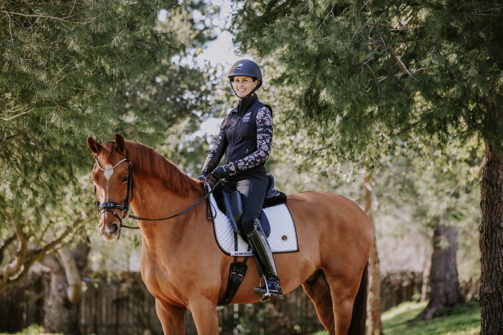 A person wearing a helmet and riding gear demonstrates excellent rider position while sitting on a brown horse in an outdoor Omaha setting surrounded by green trees and sunlight. Both appear calm and relaxed.