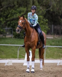 A smiling equestrian wearing a floral-patterned shirt, black helmet, and boots sits atop a chestnut horse with white leg wraps in an outdoor arena, surrounded by greenery—ready for the Training Scale Masterclass.