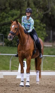 A smiling equestrian in a floral shirt and black helmet sits on a chestnut horse with white leg wraps, both ready for a Training Masterclass on the sandy arena, framed by lush green foliage in the background.
