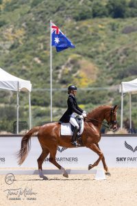A rider in formal dressage attire guides a chestnut horse in an arena, exemplifying the Training Scale, with an Australian flag, sponsor banners, and green hills in the background.