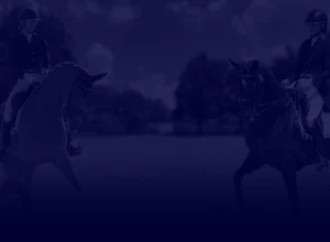 Two equestrians wearing helmets and riding attire sit on their horses, facing each other in an outdoor arena, ready for a Masterclass focused on the Training Scale. The image has a dark blue overlay with trees and a cloudy sky in the background.