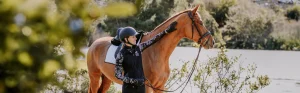 A person wearing a helmet and riding gear stands next to a brown horse, gently touching its neck, surrounded by greenery and water in the background on a sunny day—perfect for discovering free resources on horseback riding.