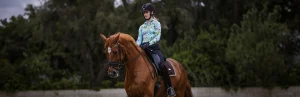 A person wearing a black helmet, colorful long-sleeve shirt, and black pants rides a chestnut horse outdoors during dressage training, with trees and greenery in the background.