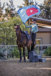 A person in riding gear stands on a mounting block next to a horse, holding a blue Amelia Newcomb Dressage flag with a YouTube logo, showcasing important Groundwork Essentials in a dirt arena surrounded by trees and a building.