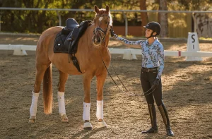A woman in riding attire stands in a sandy arena, smiling and gently patting a chestnut horse wearing a black saddle and white leg wraps. Sunlight filters through trees in the background, capturing a joyful Strides 2024 moment.