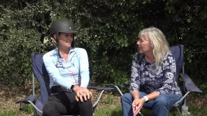Two women, one in a helmet and blue riding shirt, the other with gray hair, discuss dressage ringside. Green bushes behind them.