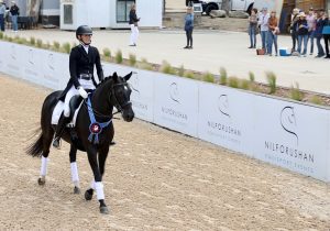 A rider in formal equestrian attire guides a black horse with a red and blue ribbon on its bridle during a dressage event, as spectators watch from the sidelines—reminding us of the focus you’ll learn at our Free Webinar. Save your Seat today!.
