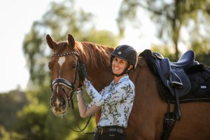 Amelia Newcomb smiling and posing with her chestnut horse who is wearing a dressage saddle and snaffle bridle.