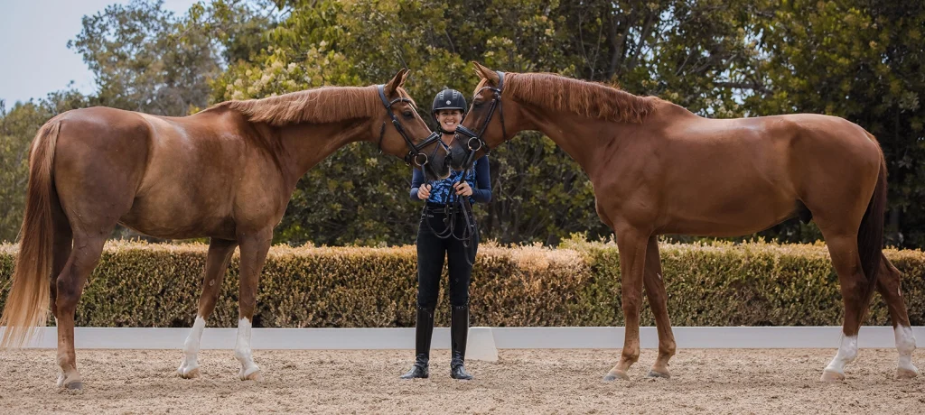 Amelia Newcomb posing and smiling with her two chestnut dressage horses, Harvey and Kensington.