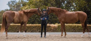 Amelia Newcomb posing and smiling with her two chestnut dressage horses, Harvey and Kensington.