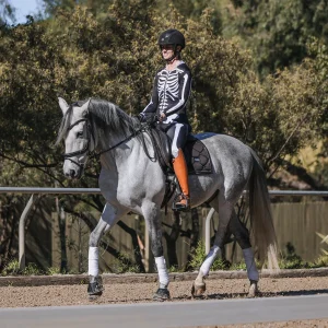 A person wearing an equestrian helmet and a skeleton costume takes on the Sitting Trot Challenge, riding a grey horse with white leg wraps and orange boots along an outdoor path, surrounded by trees in the background.