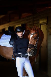 A person in equestrian attire, including a black helmet and white pants, stands smiling beside a brown horse with a white saddle pad inside a stable, ready to improve their rider fitness and master the perfect sitting trot.