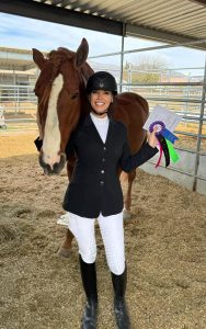 Esther Nava wearing show attire, smiling and holding a rosette while posing with her horse.