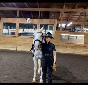 Linda, a Strides member, stands indoors next to a white horse in riding gear and helmet, smiling inside an arena with wooden walls and a large window. Riding arena letters B and P are visible on the wall behind them.