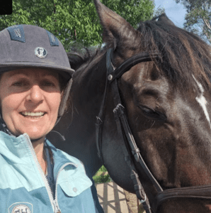 A woman in a helmet and light blue jacket smiles beside a dark brown horse with a bridle, outdoors on a sunny day with trees behind them—perfect for the 2024 Riding Bootcamp experience.