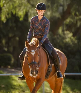 A person wearing a helmet, patterned long-sleeve shirt, and riding pants is smiling while riding a chestnut horse outdoors on a sunny 2024 day. The scene highlights a joyful moment inspired by the principles of the Training Scale. Trees blur in the background.