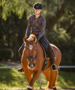 A person wearing a helmet and patterned long-sleeve shirt is smiling while riding a brown horse outdoors, with sunlight and greenery in the background, enjoying the freedom of 30DTR.