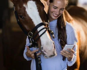 A smiling woman in a blue shirt holds a 3.0 V2 smartphone while standing beside a brown horse with a white stripe on its face. Both appear to be looking at the phone together inside a stable, capturing their bond with her 30DTR.