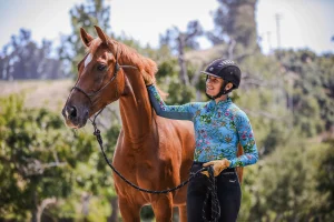 A person wearing a helmet and floral long-sleeve shirt smiles while gently holding and petting a brown horse outdoors, surrounded by trees and greenery—discover more moments like this in our 2024 Update with Free Resources.