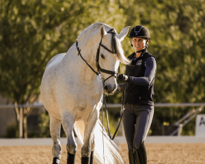 A smiling equestrian in riding gear leads a white horse wearing a bridle and leg wraps in a sandy outdoor arena, with trees and sunlight in the background—much like embracing a 30 day challenge to improve golf skills through dedication and practice.