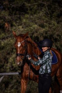 A woman wearing a helmet and a patterned shirt stands beside a brown horse with a blue saddle, smiling and patting its neck during her 30 Days to Round journey. Trees and lush greenery form the scenic background.