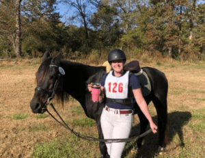 A smiling equestrian in white riding pants and helmet stands outdoors holding a drink, one hand on the reins of a black horse. The rider’s shirt has the number 126. Trees and grass fill the background—perfect after attending a Flying Changes Webinar.