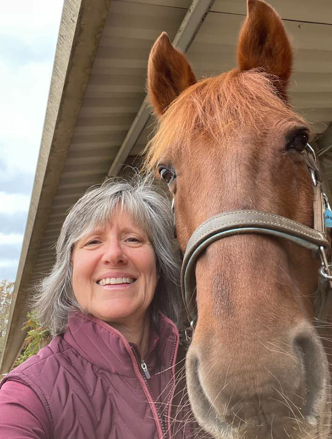 A smiling woman with gray hair takes a close-up selfie with a brown horse wearing a bridle, both under a covered outdoor structure, showcasing the groundwork of their bond.