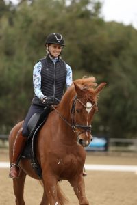 Amelia, smiling in a riding helmet and vest, is enjoying winter riding on a chestnut horse in an outdoor arena, with trees visible in the background.