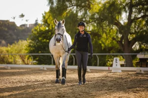 Amelia, in equestrian gear, walks beside a gray horse in a sandy outdoor arena surrounded by trees and sunlight. Dressage markers and essentials to piaffe are visible in the background.