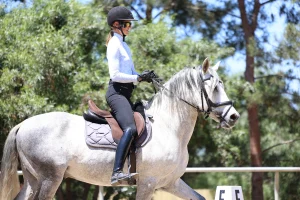 A person wearing a helmet and riding gear demonstrates excellent Rider Position while riding a white horse in an outdoor arena surrounded by trees. The rider holds the reins and appears focused, following their Rider Position Blueprint.