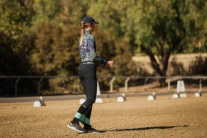 A woman in a cap, patterned long-sleeve shirt, black pants, and boots stands on a sandy arena outdoors, with trees and dressage markers in the background—showcasing ideal rider position as part of her riding technique.