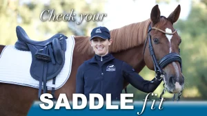 A smiling woman in a black jacket and cap stands next to a brown horse wearing a bridle and white saddle pad. Text reads, Assess saddle fit. The background is outdoors with blurred greenery.