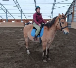A person wearing a helmet and maroon jacket is smiling with confidence while sitting on a light brown horse with a blue saddle pad inside a spacious indoor riding arena—perfect for trying out new confidence tips.