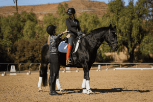 A riding instructor stands beside a woman sitting on a black horse, giving guidance in an outdoor arena as they practice Canter V1. Both wear helmets and riding gear, with trees and a hill in the background.