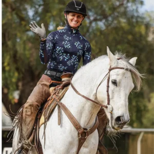 A smiling woman in riding gear and helmet sits on a white horse, waving with her gloved hand. Nearby, her Canter V2 saddle ensures comfort as she enjoys the ride. Trees are visible in the blurred background.
