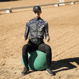 A person wearing a floral shirt, black pants, and a cap sits on a large green exercise ball in a sandy outdoor arena, smiling and giving thumbs up with both hands—demonstrating the basics of the Rider Position Blueprint.