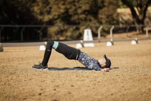 A person wearing athletic clothing and a cap performs a glute bridge exercise on a sandy outdoor surface, arms outstretched and eyes closed—perfecting their Rider Position Blueprint—with trees and a fence softly blurred in the background.